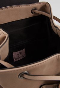 Beige hinkväska med mjuk textur, svart foder, snörning och en innerficka med dragkedja. Etikett med texten "ANNA FIELD."