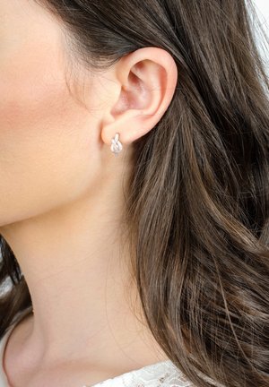 Silberne Steckerohrringe in Form von ineinander verschlungenen Schlaufen, die kleine klare Steine für zusätzlichen Glanz besitzen. Getragen von einem Modell mit langen, dunklen Haaren.