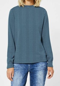 Långärmad blus i teal-färgad tyg med ett fiskbensmönster, hög halsringning och avslappnad passform, kombinerad med ljusblå denimjeans.