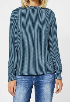 Blouse à manches longues en tissu bleu canard avec un motif en chevrons, col haut et coupe décontractée, associée à un jean en denim bleu clair.