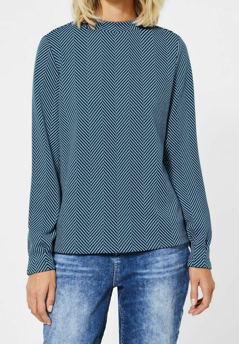 Långärmad blus i teal-färgad tyg med ett fiskbensmönster, hög halsringning och avslappnad passform, kombinerad med ljusblå denimjeans.