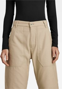 Béžové kalhoty s širokým střihem a vysokým pasem, se předními kapsami a viditelnými švy, v kombinaci s černým pleteným topem s dlouhým rukávem.