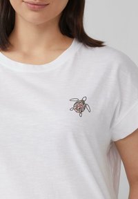 Witte korte mouwen t-shirt met een ronde, veelkleurige schildpadborduring op de linkerkant van de borst. Zacht, lichtgewicht stof.