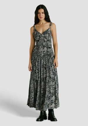 Donna con capelli lunghi e scuri che indossa un vestito lungo a fantasia bianco e nero con spalline annodate, abbinato a stivaletti neri, in piedi su uno sfondo grigio.