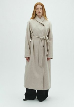 Donna con capelli rossi indossa un cappotto lungo beige con cintura sopra pantaloni neri a gamba larga, in piedi davanti a uno sfondo chiaro uniforme.