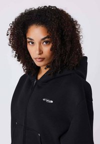 Sudadera negra con cremallera y capucha grande, con bolsillo delantero y un pequeño logo plateado en el pecho. Tela suave y texturizada.