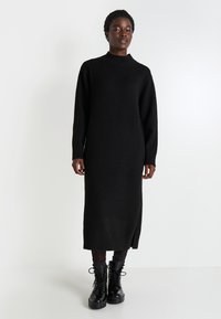 Robe tricotée noire avec un col montant, des manches longues et une fente sur le côté à l'ourlet. Associée à des bottes de combat noires à lacets, au toucher lisse.