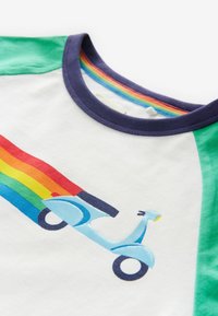 Balta kokvilnas T-krekls ar zaļām piedurknēm un zilu skūteri attēlu ar varavīksnes pēdas zīmējumu. Tumši zils ribota apkakle.