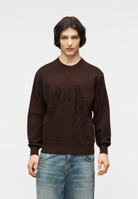 Νέος με κοντά σκούρα μαλλιά, φορώντας ένα καφέ φούτερ με μαύρο αφηρημένο σχέδιο και φαρδιά μπλε τζιν, μπροστά σε απλό φόντο.