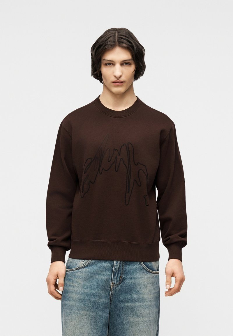 Νέος με κοντά σκούρα μαλλιά, φορώντας ένα καφέ φούτερ με μαύρο αφηρημένο σχέδιο και φαρδιά μπλε τζιν, μπροστά σε απλό φόντο.