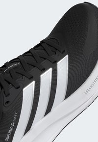 Black athletic shoe with textured upper, featuring three white stripes, a cushioned sole, and a streamlined design. "SUPERNOA EASE 2" branding visible.