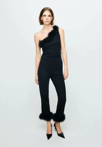 Combinaison noire avec une épaule dénudée et une bordure en fausse fourrure le long de l'encolure et des poignets. Coupe ajustée, texture lisse, portée avec des talons aiguilles.