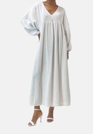 Kobieta ubrana w długą, luźną białą sukienkę z bufkowymi rękawami i dekoltem w serek, do tego białe sandały na obcasie z paskiem wokół kostki.