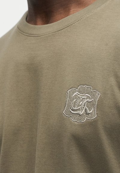 Κοντινό πλάνο πράσινου ελιάς t-shirt με κεντητό λευκό περίτεχνο λογότυπο στην αριστερή πλευρά του στήθους.