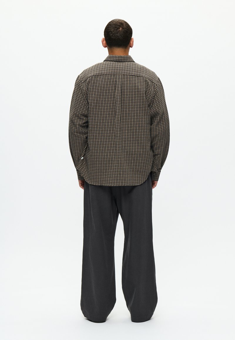 Camicia a quadri marrone e nera, con apertura a bottoni e polsini ripiegati, abbinata a pantaloni grigi larghi. Tessuto testurizzato.