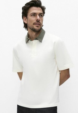 Mężczyzna o ciemnych włosach ubrany w białą koszulkę polo z krótkim rękawem i zielonym kołnierzykiem, stoi z rękami za plecami na jednolitym tle.