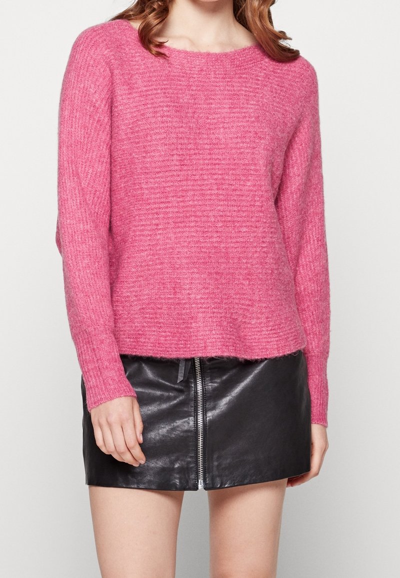 Femme portant un pull rose duveteux et une mini-jupe en cuir noire avec une fermeture éclair à l'avant, debout devant un fond clair uni.
