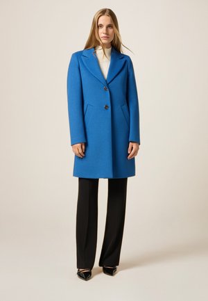 Cappotto di lana blu con un design strutturato, bottoni doppi e tasche laterali. Abbinato a pantaloni neri e scarpe con punta.