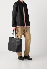 Czarna, lekka, wodoodporna kurtka z kołnierzem i sznurkiem. Jasnoróżowy T-shirt pod beżowymi spodniami. Czarna skórzana torba na ramię z wytłoczonym napisem "HUGO."