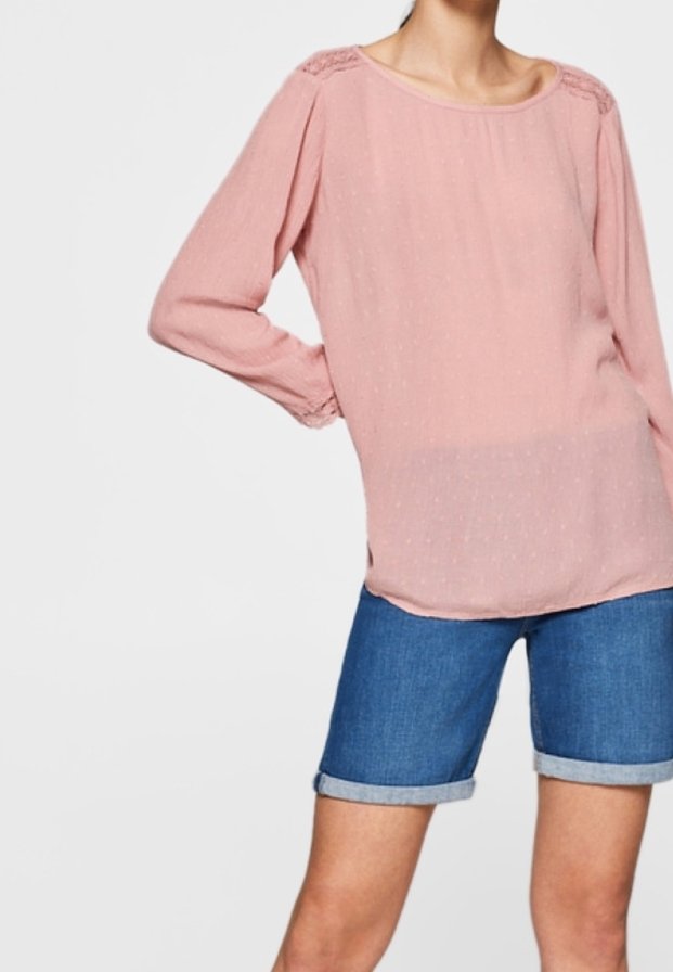Rosa Bluse mit langen Ärmeln, Rundhalsausschnitt und strukturierten Punkten, kombiniert mit blauen Denim-Shorts, die am Saum umgekrempelt sind. Strukturierte Akzente an den Schultern.