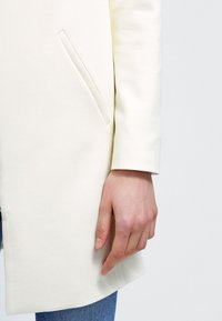 Vit kappa med slim fit, slät textur och sidoficka. Ärmen har en ren finish som visar minimalistiska designdetaljer.