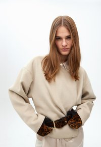 Beige fleece-pulloweri, jossa on kaulus ja nappidetalji, yhdistettynä mustaan vyöhön, jossa on leopardikuvio ja kultaiset yksityiskohdat.