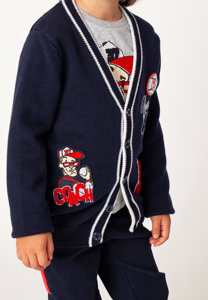 Marineblå strikket cardigan med hvid kant, der har broderede mærker af en træner og en baseball. Knapper foran; lommer med dekorative detaljer.