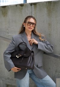 Šedý blazer, světle modré džíny, tmavě hnědá kožená kabelka se dvěma uchy a zlatými kovovými detaily. Hladký povrch, strukturovaný design.