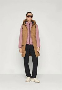 Brown quilted vest with a hood over a pink zip-up top and black pants. Sunglasses and light-colored sneakers complete the outfit.