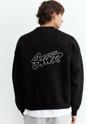 Personne aux cheveux courts et bruns de dos, portant un sweat-shirt noir avec un texte blanc stylisé dans le dos, par-dessus une chemise blanche et un pantalon gris.