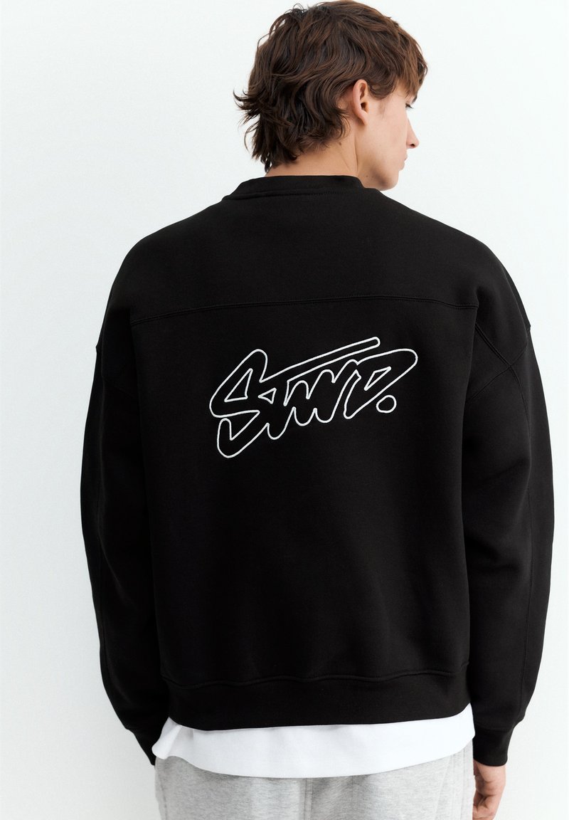 Personne aux cheveux courts et bruns de dos, portant un sweat-shirt noir avec un texte blanc stylisé dans le dos, par-dessus une chemise blanche et un pantalon gris.
