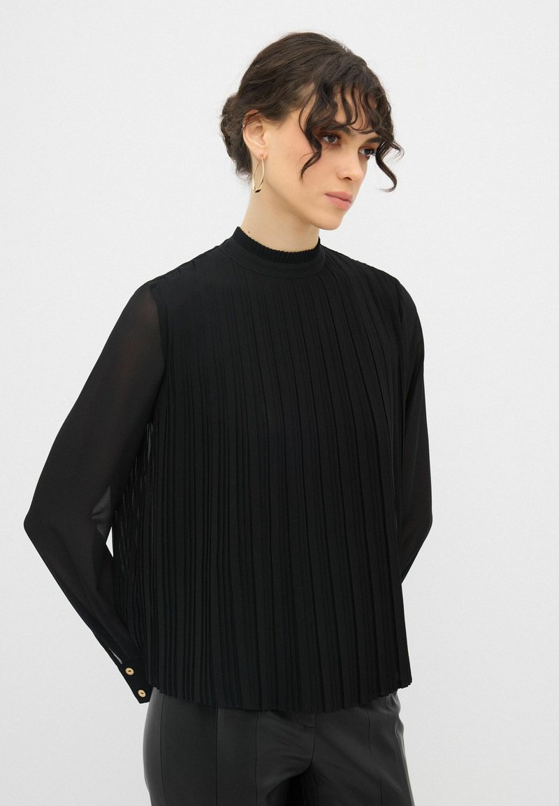 Blusa nera plissettata con maniche lunghe trasparenti, collo alto e dettagli con bottoni dorati ai polsini. Tessuto liscio, silhouette moderna.