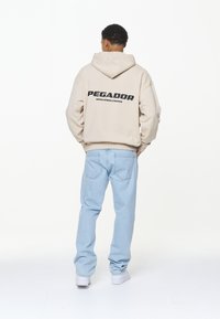 Beige Kapuzenpullover mit großem schwarzen "PEGADOR"-Text auf dem Rücken, kombiniert mit hellblauen Jeans und weißen Sneakers, von hinten betrachtet.