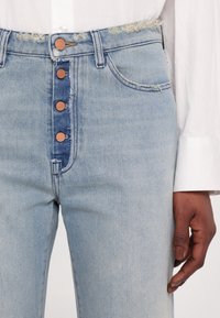 Lyseblå denimjeans med høj talje, fem kobberknapper, frynsede kanter og to forlommer. Glat tekstur og falmet finish.