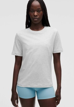 Frau mit langem geflochtenem Haar, die ein schlichtes hellgraues kurzärmliges T-Shirt und hellblaue eng anliegende Shorts vor einem schlichten Hintergrund trägt.