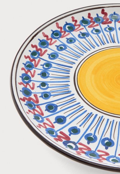 Assiette en céramique ronde avec un centre jaune, un motif floral bleu et vert, et des accents rouges en zigzag le long du bord blanc.