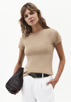 Kvinde med bølget hår iført beige kortærmet top, hvide bukser med sort bælte, der holder en stor mørk flet clutch i venstre hånd.