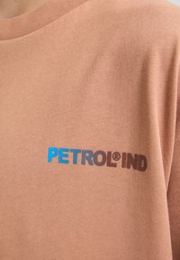 Camiseta de algodón marrón con el texto impreso en azul y marrón "PETROL® IND" en el lado izquierdo. Textura suave con cuello redondo.
