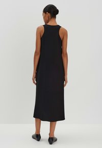 Schwarzes, ärmelloses Maxikleid aus weichem Stoff, mit slim fit, rundem Ausschnitt und einem Saum bis unter das Knie. Flaue schwarze Schuhe.