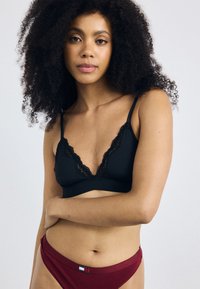 Musta pitsireunuksilla varustettu bralette, jossa on säädettävät olkaimet ja syvä V-kaula-aukko, yhdistettynä punaisiin bikineihin, joissa on merkkilogo.