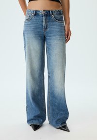 Široke modre kavbojke iz denima z zbledelim pranjjem, z visokim pasom, sprednjimi žepi in kovinskimi dodatki. Teksturovan material.