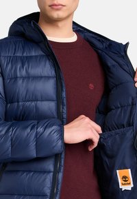 En person i en marinblå puffjacka öppnar den, vilket avslöjar en vinröd tröja under, som har en Timberland-logga på innerfickan.
