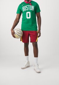 Persoon in groene "Boston 0" shirt, rode shorts met gele strepen, witte sneakers en rode bandana die een witte basketbal met het Nike-logo vasthoudt.