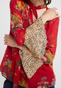 Mujer con blusa roja de flores y mangas acampanadas de estampado de leopardo, cruzando los brazos sobre el pecho.