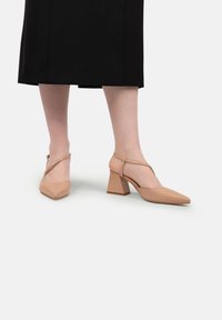 Picioare purtând pantofi cu toc bloc bej, cu vârf ascuțit și curea pentru gleznă, asortați cu o fustă midi neagră, pe un fundal simplu.