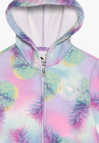 Sweat à capuche zippé dans des couleurs pastel, avec un motif d'ananas et un logo circulaire. Tissu doux avec une surface légèrement texturée.