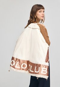 Chaqueta con capucha blanca y cobre con el texto "COLO CLUB", que presenta un acabado brillante, un dobladillo con cordón y detalles intrincados en el cuello.