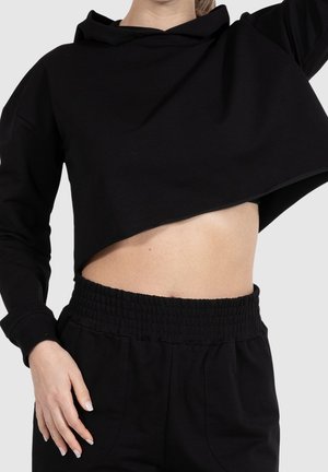 Femme portant un sweat à capuche court noir et un pantalon noir taille haute, levant le bras pour montrer le ventre sur un fond uni.
