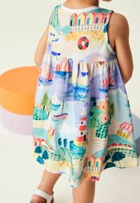 Dítě oblečené v barevných šatech s ilustracemi moře, včetně plachetnic, majáků, plážových budov, racků a stromů.