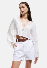 Blouse blanche cropped à manches longues, décolleté en V et lien à l'avant. Associée à un short blanc taille haute et une ceinture marron avec accent métallique.